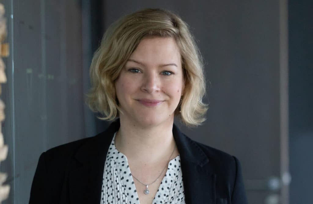 Woman with short blond hair in a black blazer, smiling indoors with a blurred background.