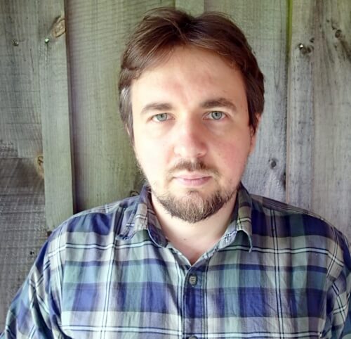 A person wearing a plaid shirt stands in front of a wooden wall, looking at the camera.