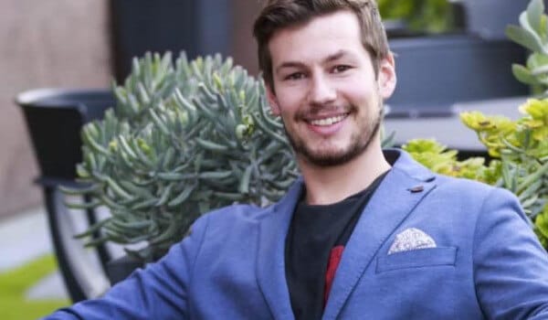 Smiling man in a blue blazer sits outdoors near green plants.