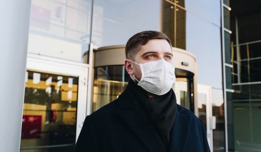 Man wearing a face mask and black coat stands outside a modern glass building.