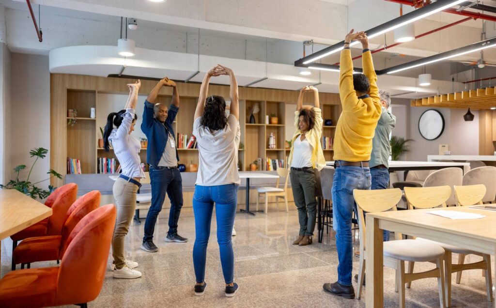 People stretching their arms upwards in a bright office space with bookshelves and tables in the background.