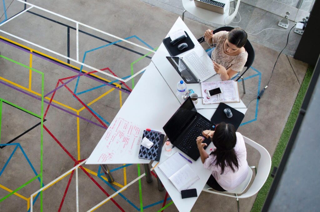 Two people working at a desk with laptops and papers, seen from above, on a floor with colorful geometric designs.
