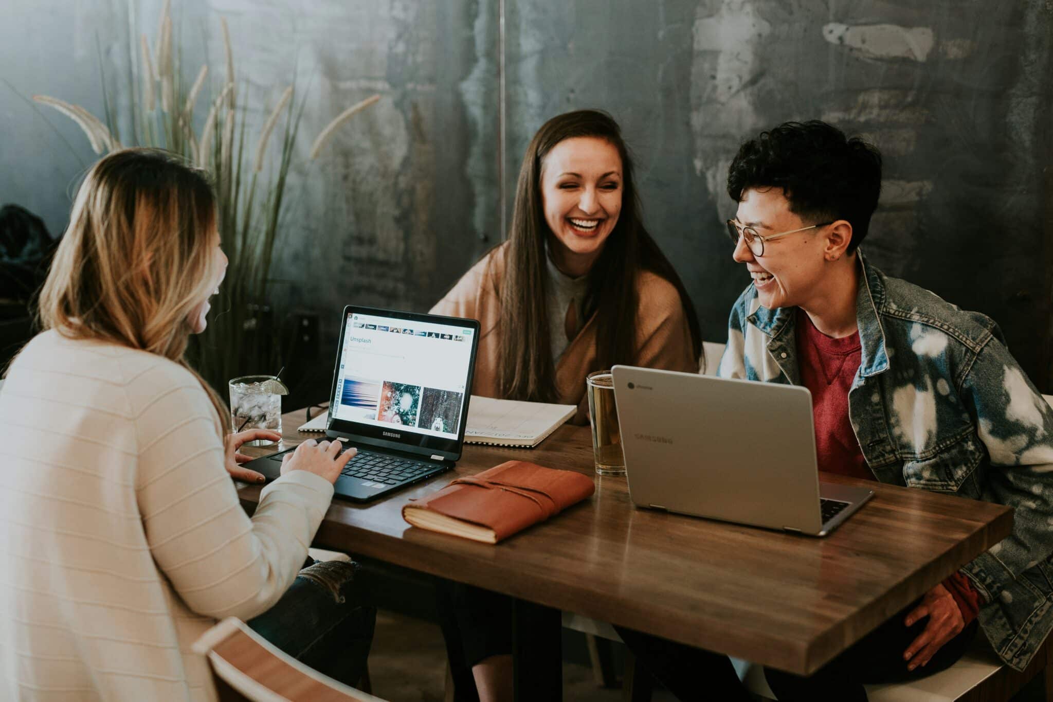 Three people laughing and working on laptops at a wooden table in a cozy cafe setting.