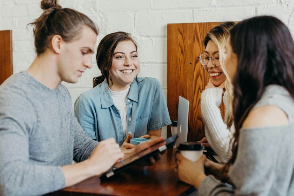 Four people are sitting at a table in a café, smiling and talking with drinks and a tablet.