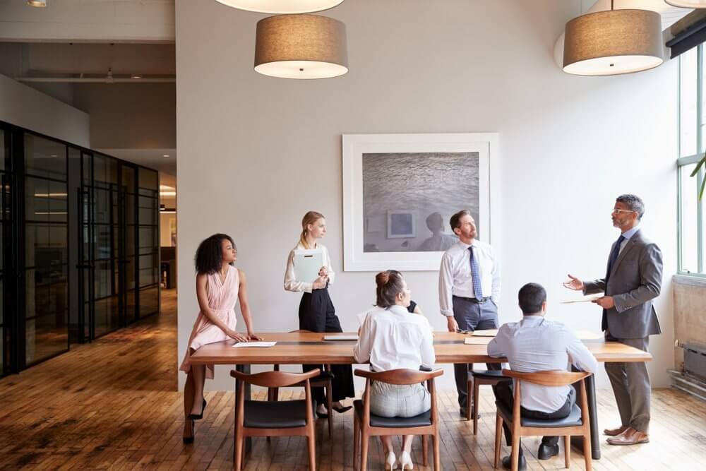 Six people in a modern office having a meeting around a long table, with one person standing and speaking.