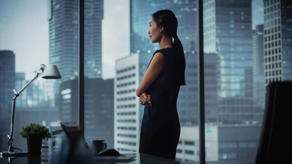 Woman in black dress stands in an office, looking out at a cityscape through large windows, with folded arms.