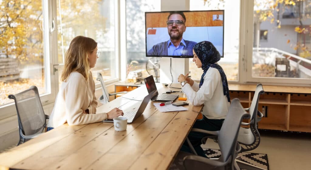 Two women at a table have a video call with a man on a screen; autumn leaves are visible through the window.