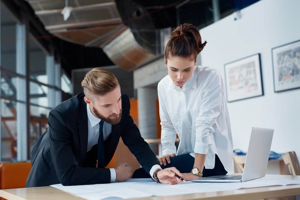 A man and woman in an office reviewing documents on a table with a laptop nearby.