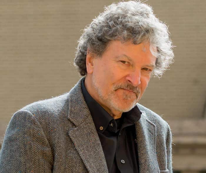 Man with curly gray hair and goatee in a gray suit jacket and black shirt, standing outdoors, with a neutral expression.