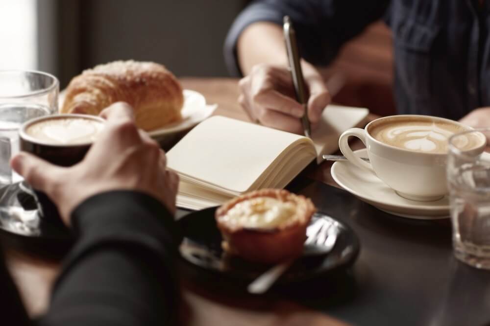Two people at a table with coffee, pastries, and a notebook. One writes while the other holds a cup.