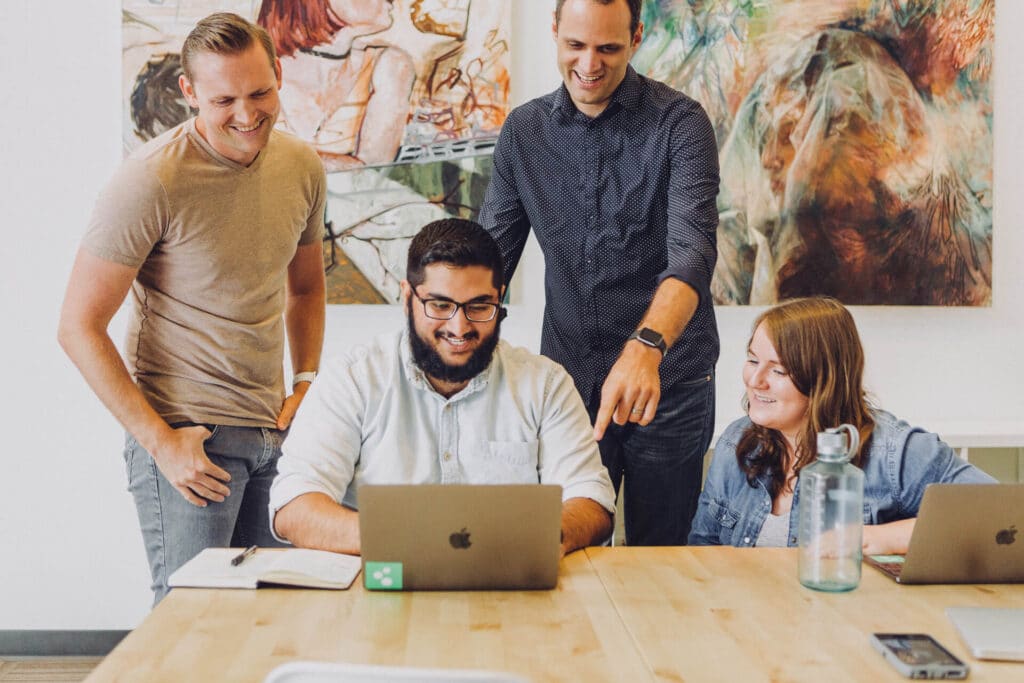 Four people happily collaborating around laptops at a table, with artwork on the wall behind them.