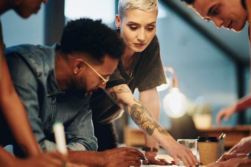 Four people collaborating around a table, one with a tattooed arm, under warm lighting.