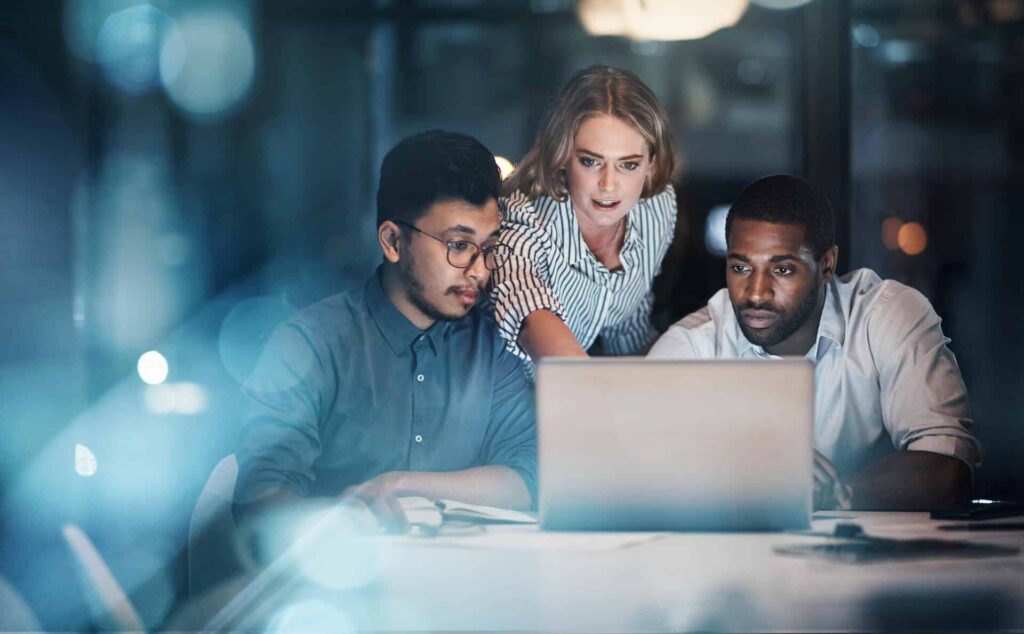 Three people intently looking at a laptop in a dimly lit office with blurry blue lights in the foreground.