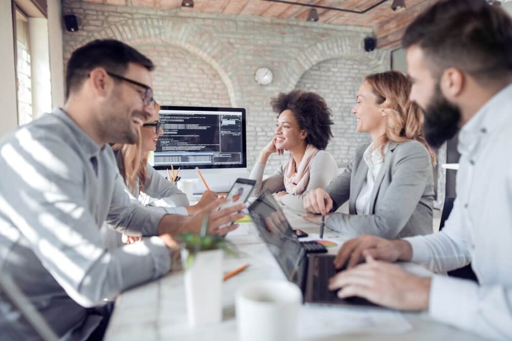 Team collaborating around a table with laptops, discussing work with a cheerful ambiance in an office setting.