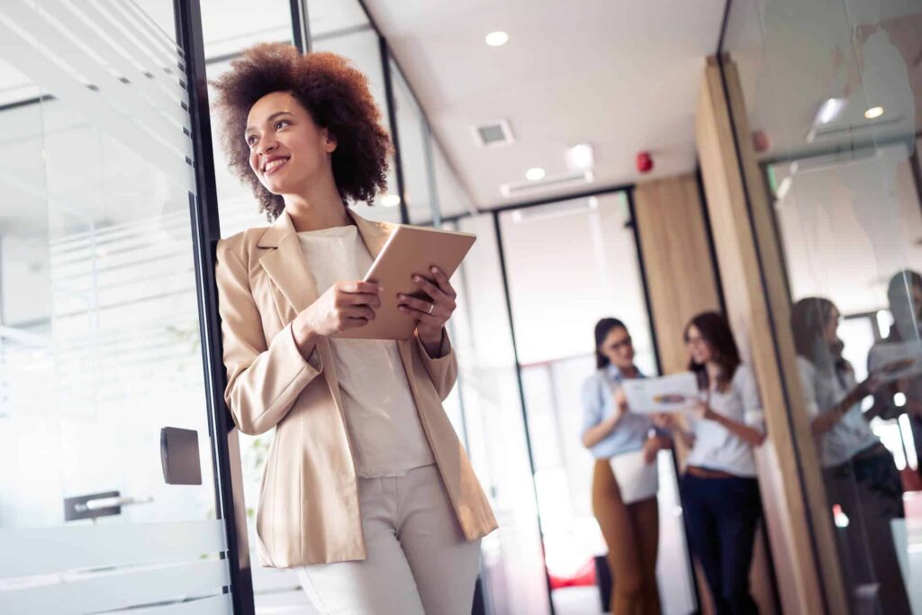 Woman in a light suit smiles while holding a tablet in a bright office corridor, with two people talking in the background.