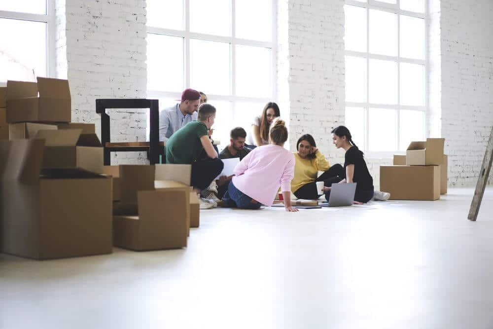 A group of people sitting on the floor in a bright room with large windows, surrounded by cardboard boxes.