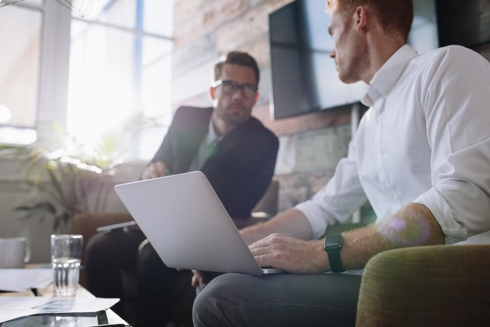Two men in an office setting are in conversation; one is using a laptop, and the other is listening attentively.