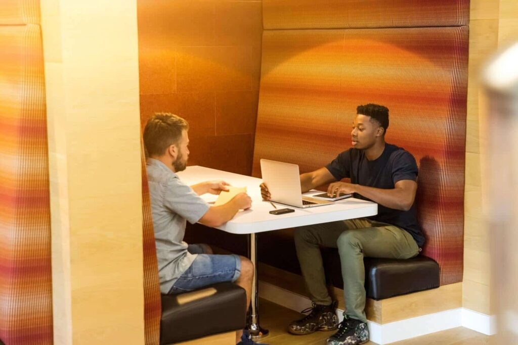 Two people sitting at a booth table with a laptop and documents in a warm-toned room.