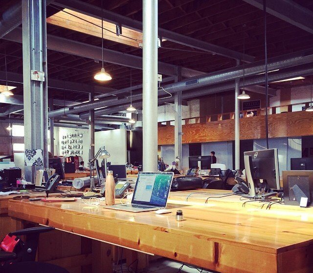 Open-plan office with wooden desks, computers, and industrial-style lighting in a spacious area.