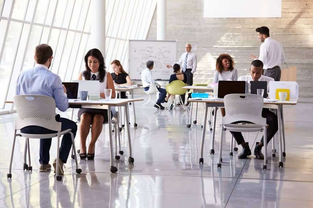 People working on laptops at tables in a bright, modern office with large windows and a meeting area.