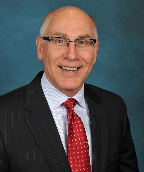 Smiling man in glasses, wearing a pinstripe suit, red tie, and white shirt against a blue background.