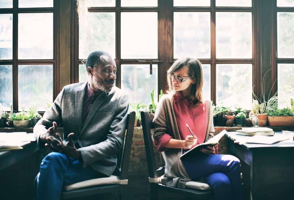 Two people sitting by a window, talking and taking notes in a well-lit room with plants on the sill.