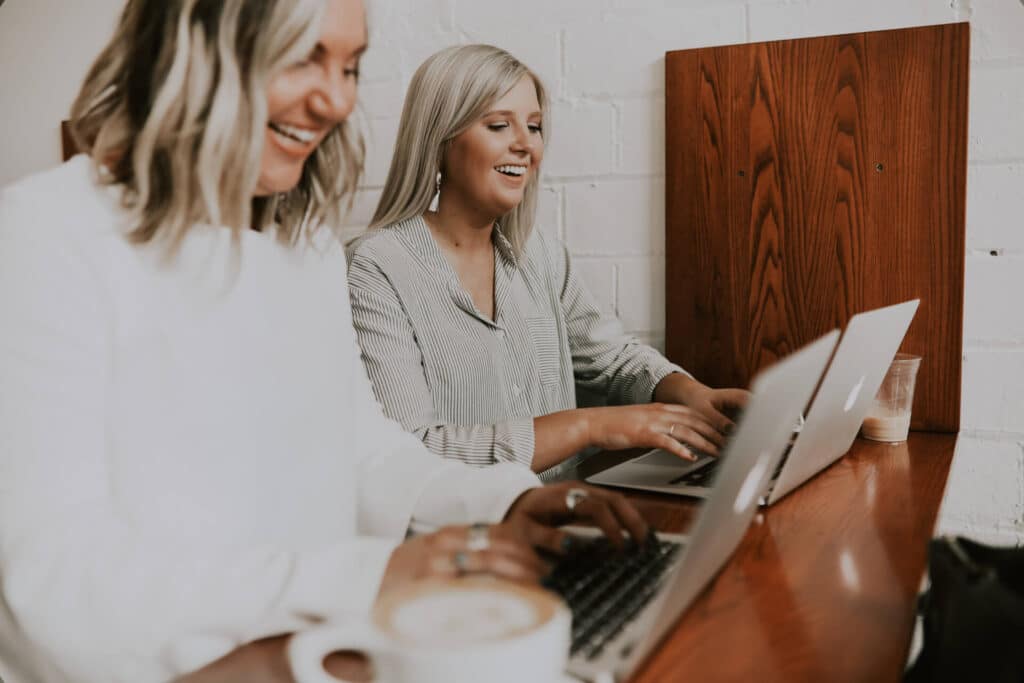 Two women smiling and typing on laptops at a wooden desk, with a cup of coffee in the foreground.