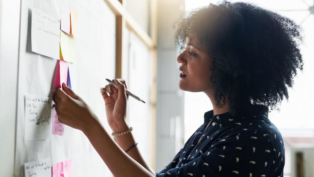 Person arranging sticky notes on a board, holding a pen, in a well-lit room.
