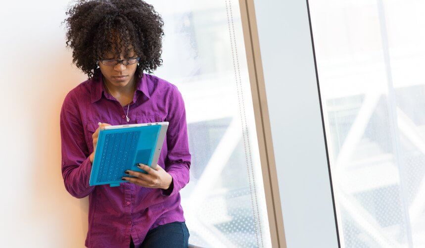 Person with curly hair, wearing glasses and a purple shirt, writes on a blue tablet near a large window.