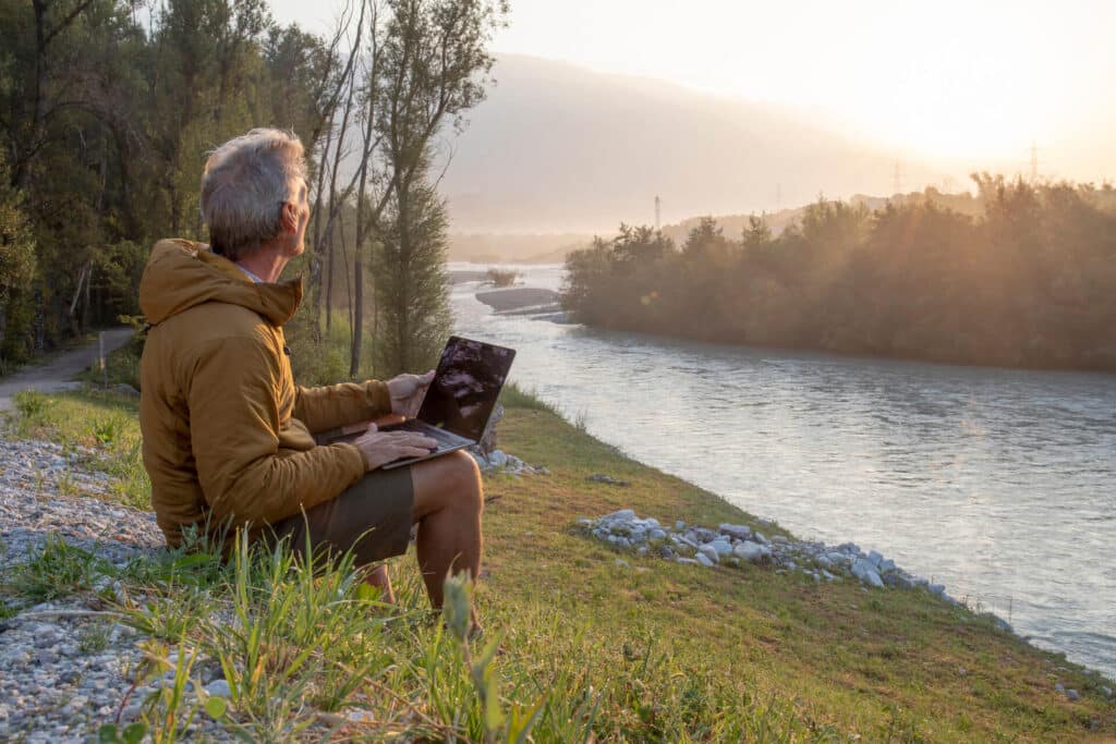 Man with laptop sitting by a riverbank, looking at a sunrise or sunset, surrounded by trees and mountains.