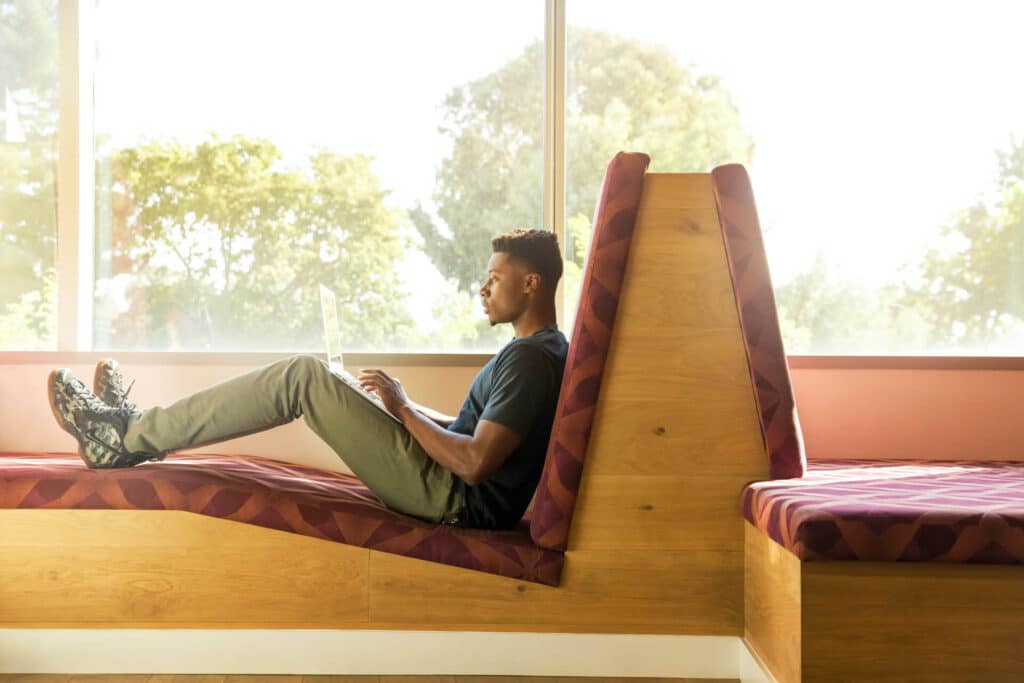 Person sitting on a cushioned wooden bench, using a laptop, with trees visible through the window behind them.