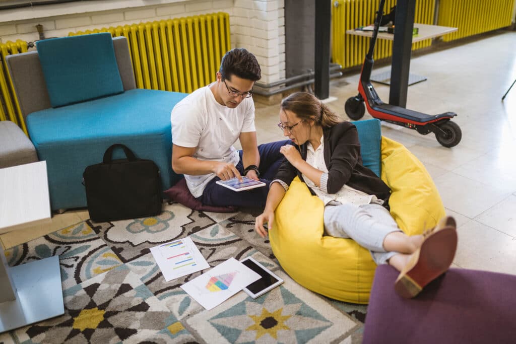 Two people sitting on colorful bean bags in an office space, discussing charts and using a tablet.