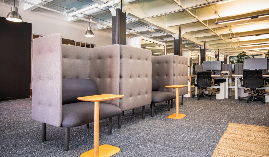 Modern office with gray partitioned seating, wooden side tables, and open desks with computers in the background.