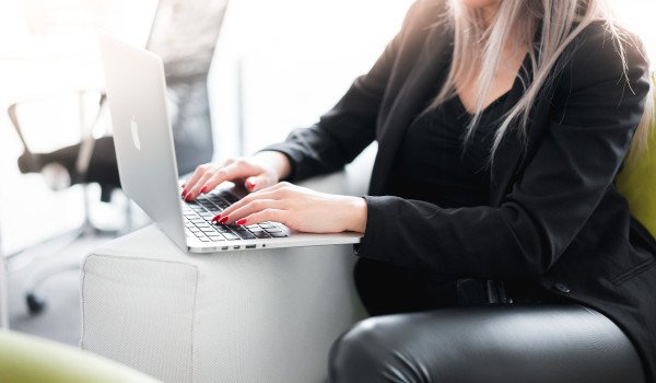 Person in black outfit typing on a laptop while sitting on a white sofa.