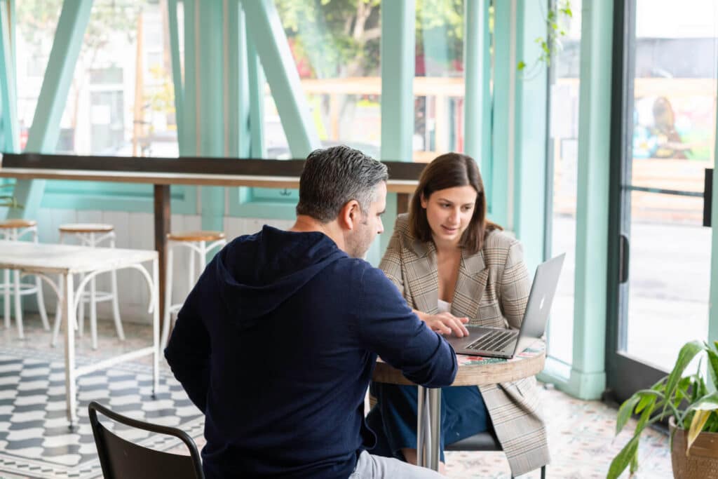 Two people at a café table, discussing and looking at a laptop, with large windows in the background.