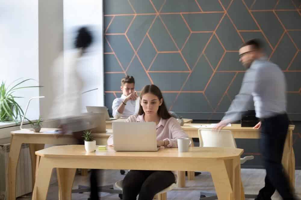 People working at desks in a modern office, with a geometric wall and blurred motion suggesting movement.