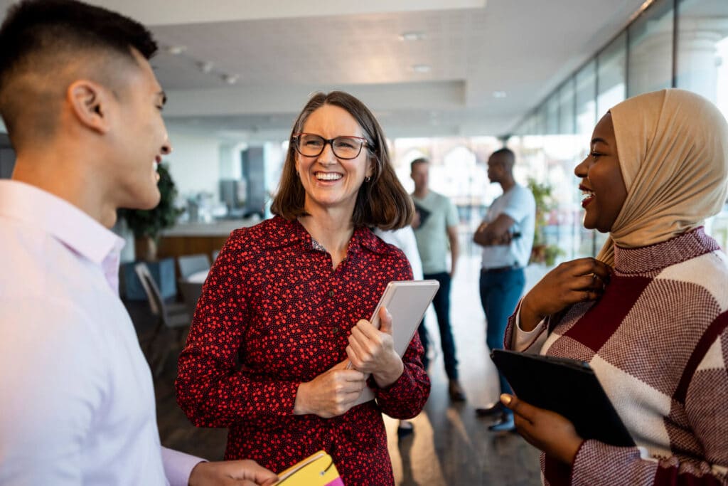 Three people are talking and laughing in an office setting. Others are in the background.