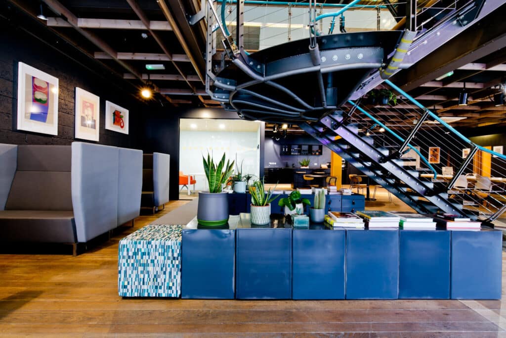 Modern office interior with a spiral staircase, plants, blue seating, and framed artwork on the walls.