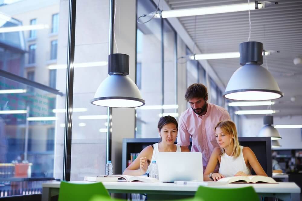 Three people studying together in a modern library, focused on a laptop, with books and water bottles on the table.