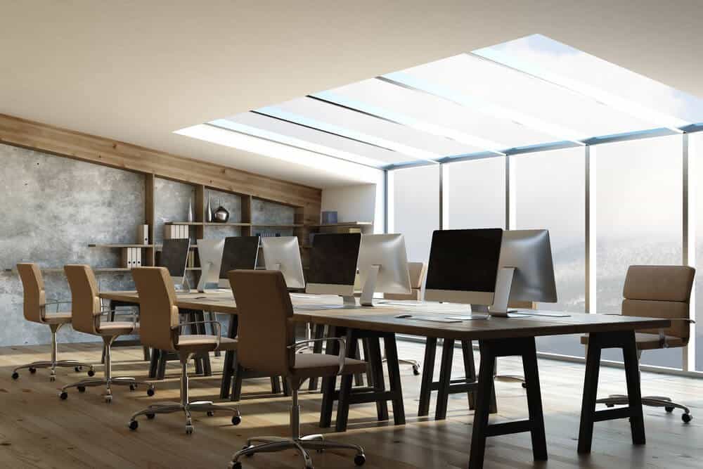 Modern office with wooden desks, beige chairs, computers, and large skylight windows.