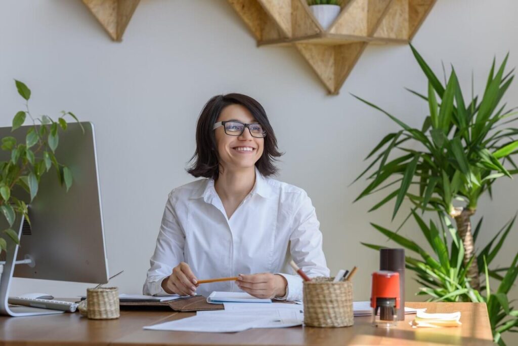 Smiling person with glasses at a desk in an office, surrounded by plants and triangular shelves.