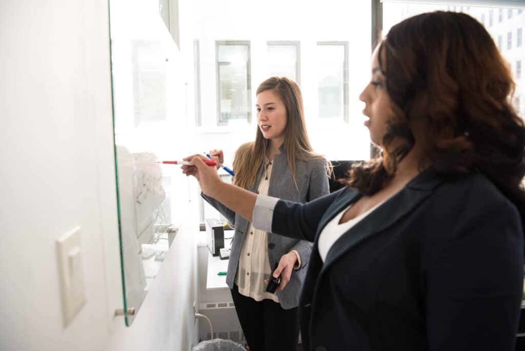 Two women writing on a whiteboard in an office setting, with one holding a marker and the other observing attentively.