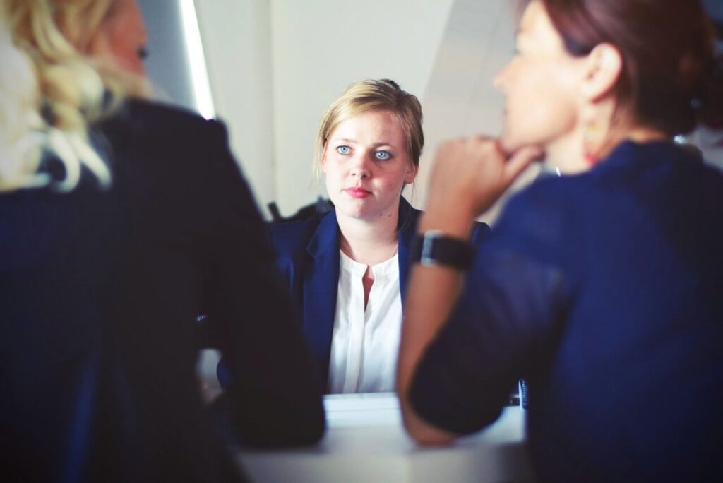 Three women in business attire having a meeting at a table. One woman is facing the camera, listening attentively.