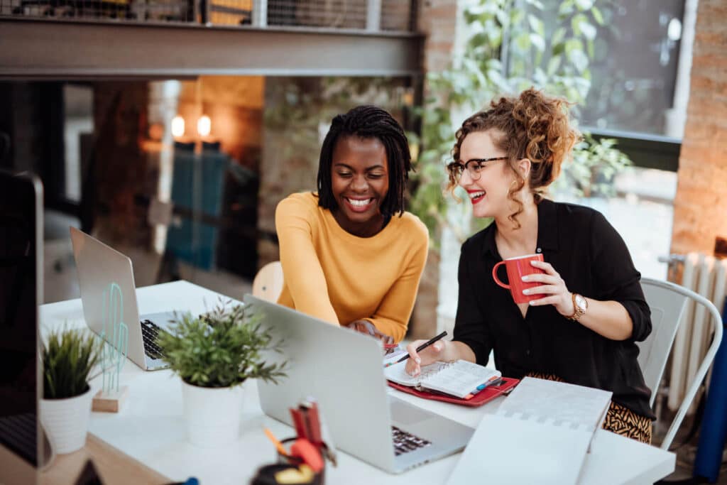 Two women are smiling while working together at a table with laptops, notebooks, and plants. One is holding a red mug.