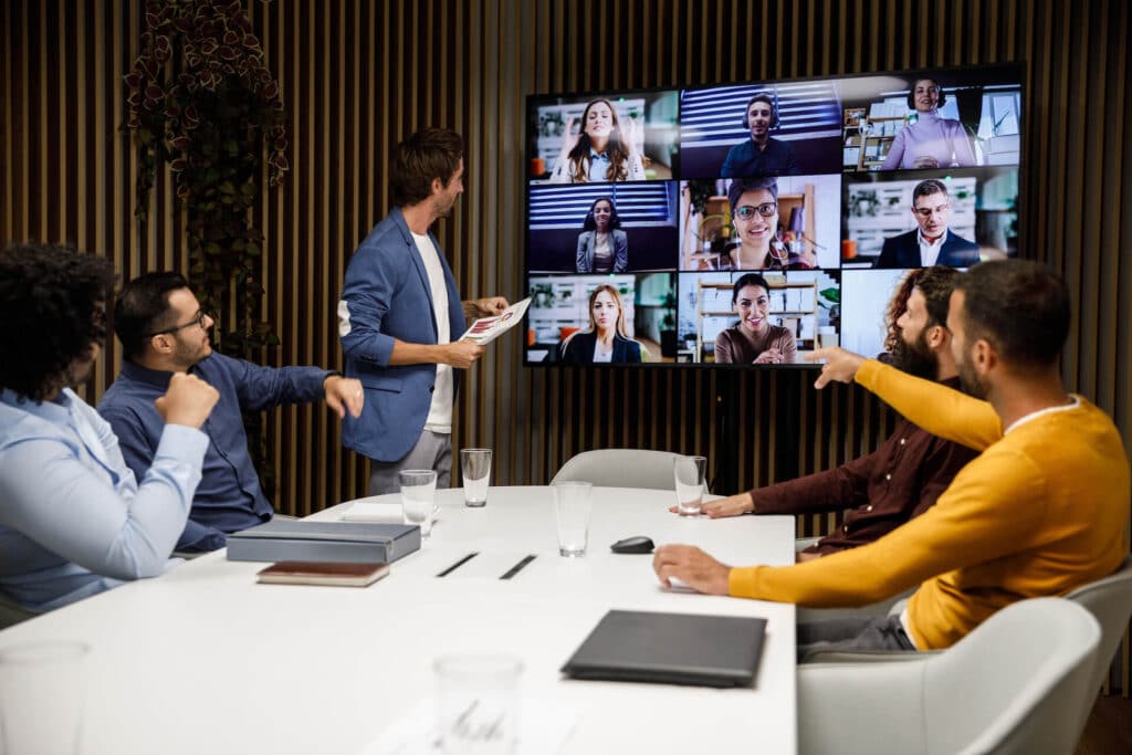 A group of people in a meeting room watching a video conference on a large screen.