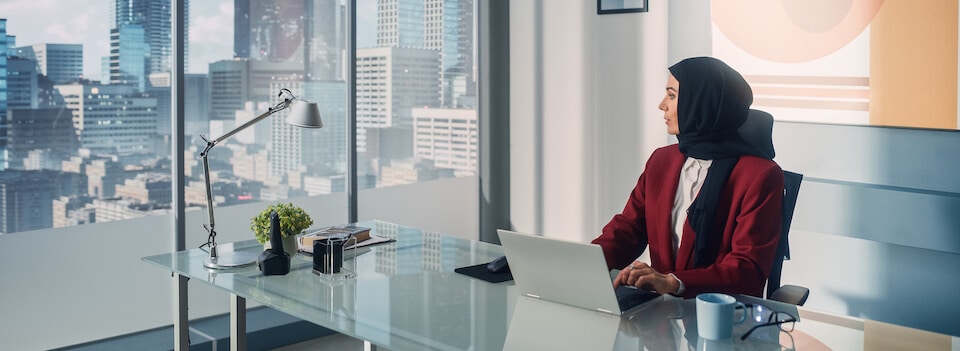 A woman in a hijab, seated at a desk with a laptop, looks out a window with a cityscape view.