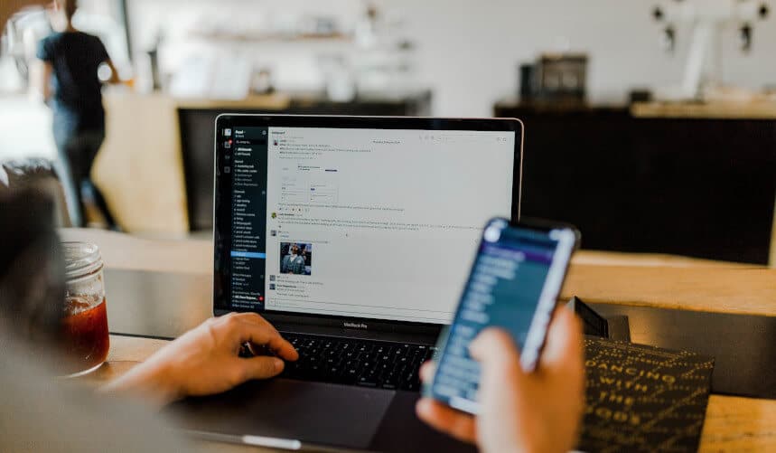 Person using a laptop and smartphone at a desk, with a blurred figure in the background.