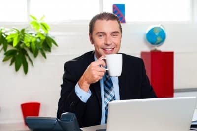 Smiling man in a suit drinking coffee at a desk with a laptop and office accessories.