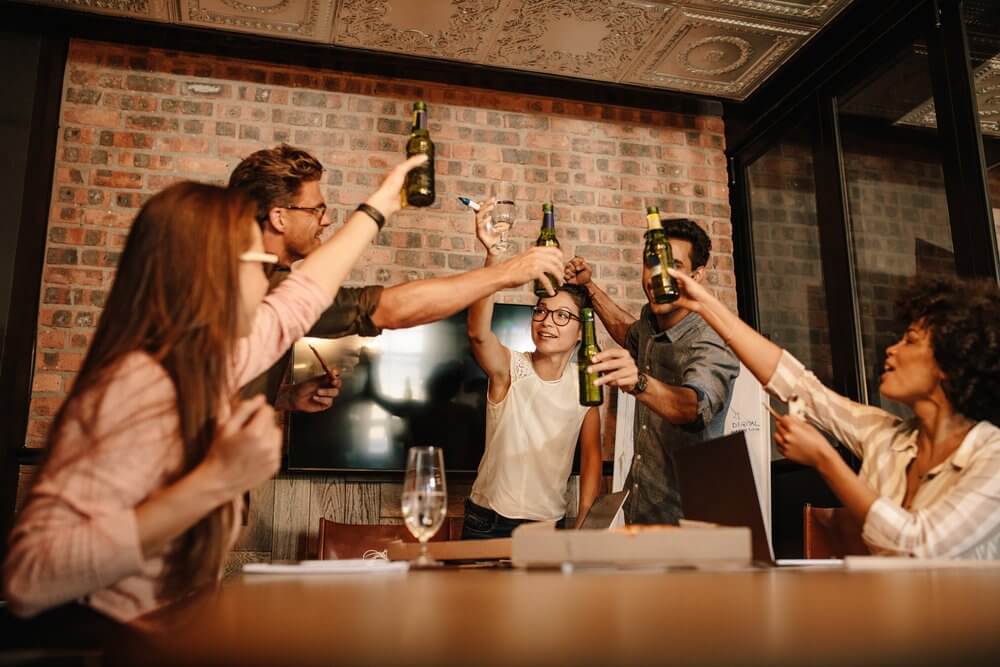 People in an office raising beer bottles and glasses in a celebratory toast around a table with a pizza box.