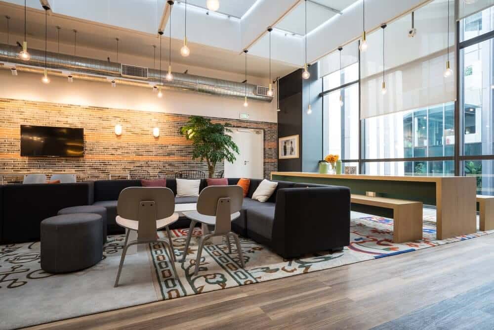 Modern lounge with sofas, tables, decorative rugs, and hanging lights. Large windows and a brick accent wall present.